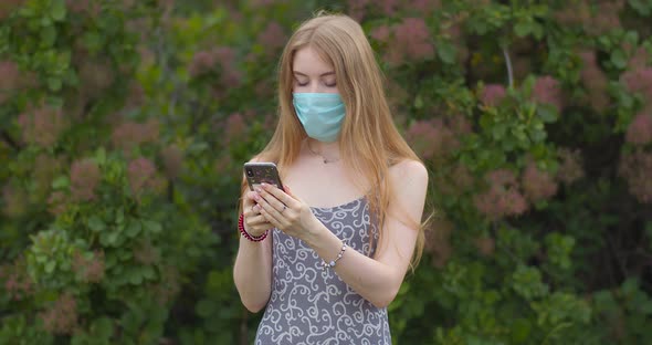 A young girl in a protective medical mask looks at the phone and sends messages