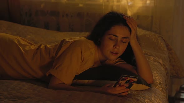 Smiling pretty young woman is lying on the bed, leaning on her arm, and using a smartphone. alt