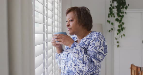 Happy african american senior woman enjoying drinking cup of tea looking out of window alt