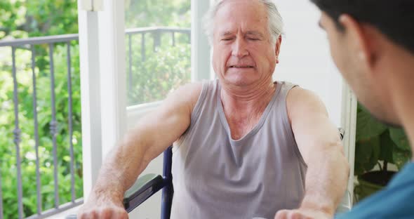 Disabled senior man grimacing while lifting dumbbells with support of male instructor at health club alt