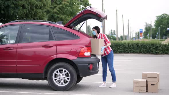 Female Courier, in Protective Mask, Gloves, Loading Parcels, Putting Cardboard Boxes To Car Trunk alt