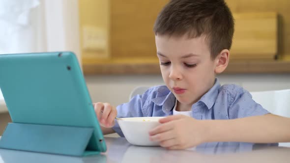 Little Boy Eating Cereal with Milk in the Kitchen and Watching Cartoon on Tablet Computer