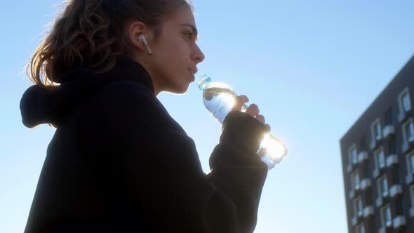 Young Woman Sits on Bench in Apartment Complex in Morning After Jogging and Drinks Water alt