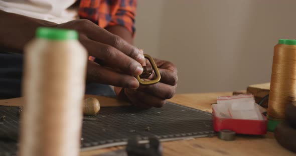Close up of hands of african american craftsman preparing belt in leather workshop alt