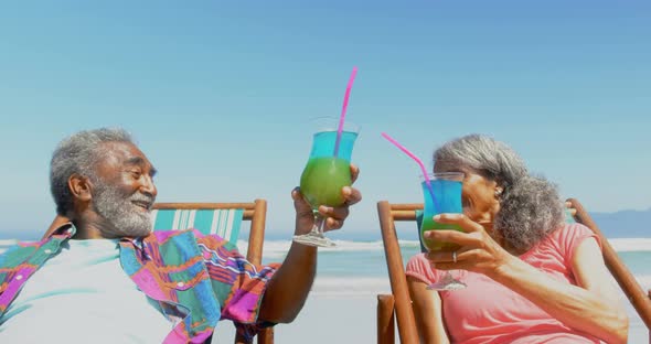 Front view of active senior African American couple toasting drinks on deckchair on the beach 4k alt