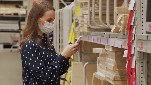 Woman Wearing a Protective Mask in Store Chooses Timber for Carpentry Work Uses Smartphone alt