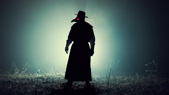 Silhouette of Scary Plague Doctor Man in Long Mantle Costume Standing in Front of Bright Spotlight alt
