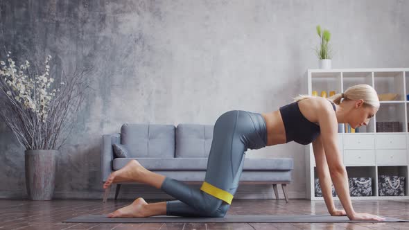 Young and sporty girl in sportswear is doing exercises in home interior using resistance band. alt