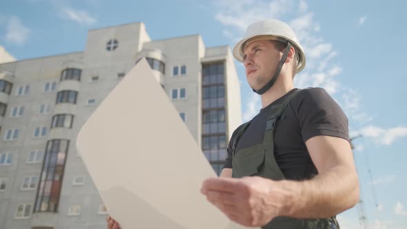 A Man in a Helmet Is Holding a Construction Plan in His Hands. A Builder Is Checking Blueprints alt