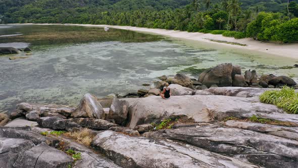 Aerial View of Blonde Tanned Girl Sitting in Yoga Pose and Meditating on Granite Rock Against Exotic