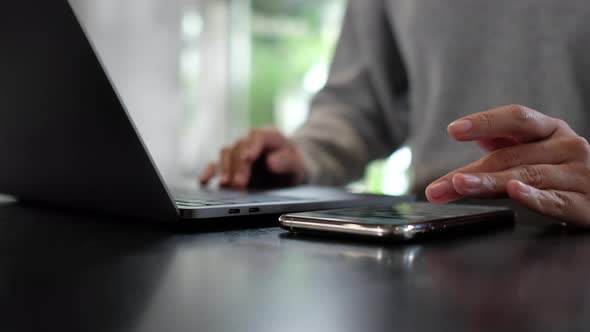 Closeup of a woman touching and scrolling on smart phone screen while working on laptop computer