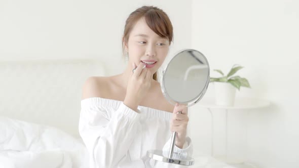 Beautiful young asian woman at the mirror holding and looking a makeup lipstick. alt