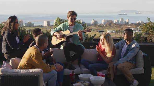 Young Caucasian man playing guitar on a rooftop with his friends alt