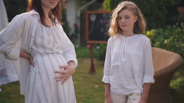 Portrait of Unsatisfied Caucasian Teenage Girl Standing with Unrecognizable Pregnant Mother on alt