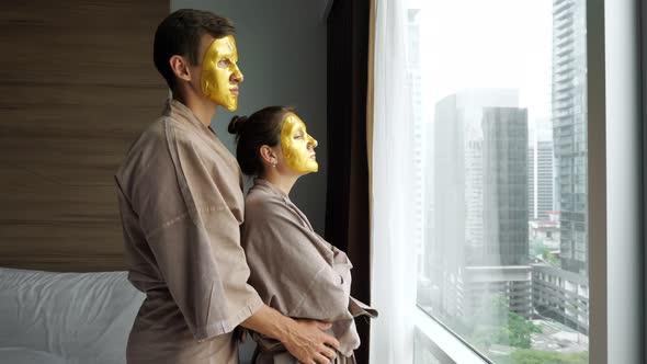 Man Hugs Girl with Golden Face Mask Near Window in Hotel alt
