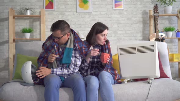 Frozen Young Woman and Man Wrapped in a Blanket in the Living Room Are Heated Next To Electric alt