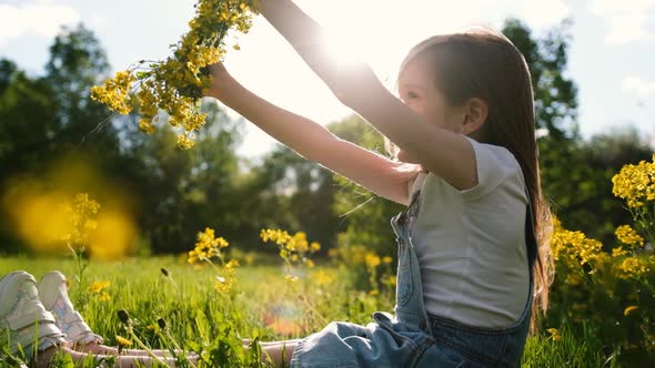 Little Cute Girl Sits on a Sunny Meadow and Twirls a Wreath of Yellow Flowers in Her Hands alt
