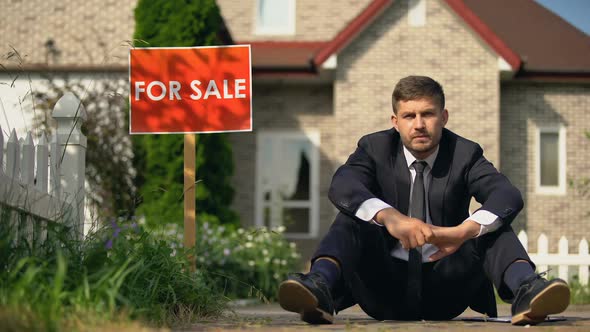 Depressed Man Sitting on Ground Near for Sale Sign, Home Loss Because of Debts alt