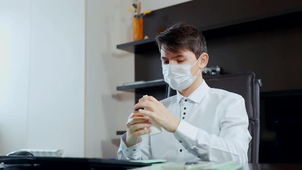 A Young Man in a Medical Mask Sitting at the Table Disinfects His Hands alt
