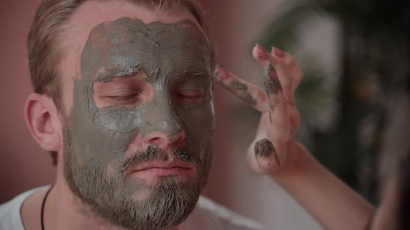 Portrait of Male Face Being Covered with Clay Mask By Cosmetician in Spa. Woman Applying Skincare