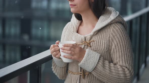 Caucasian Woman Stays on Balcony During Snowfall with Cup of Hot Coffee or Tea alt