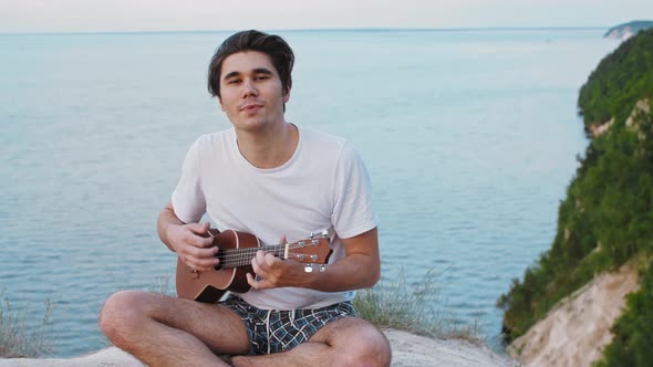 Young Smiling Man Playing Ukulele and Singing  Sitting on the Mountain and Looking in the Camera alt