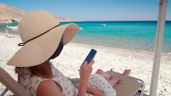 COVID-19 Tourism: Woman in Face Mask Uses Phone App on Sea Beach in Summer