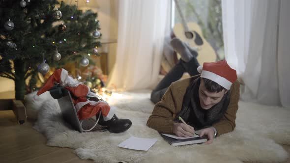 Wide Shot Portrait of Joyful Young Caucasian Man Writing Letter To Santa at Home alt