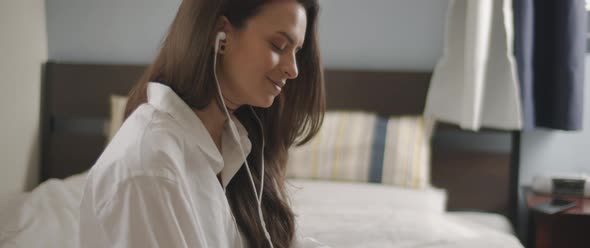 Close up of a young woman talking to someone through earphones while sitting on bed.  alt