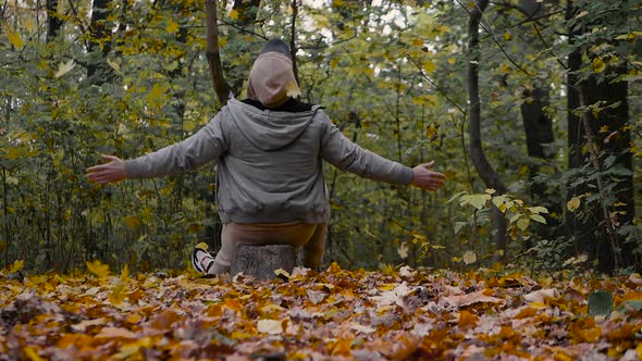 Happy Young Man Enjoys Leaf Fall In The Forest In Autumn, The Man Sits On A Stump. alt