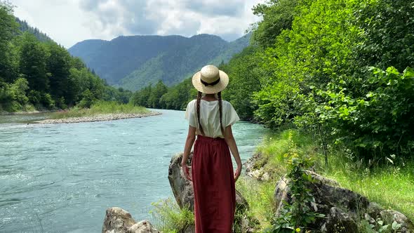 Young beautiful romantic vintage woman in straw hat going to stone coast near mountain river, travel