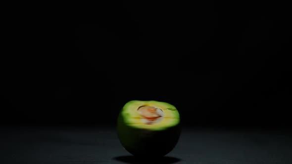 Female Hand Spinning Halved Avocado at Black Background on Table Leaving alt