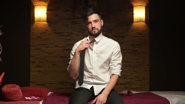 Young tired manager sits on a couch in a massage parlor and idly removes his tie alt