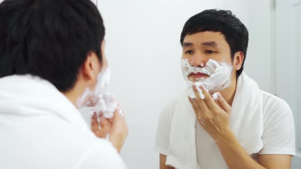 young man applying foam cream on face before shaving in the bathroom mirror alt