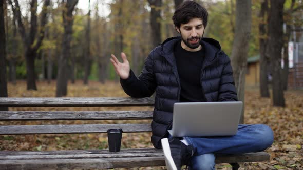 Smiling Man Sitting Casually on Bench in Public Park Using Laptop for Video Call alt