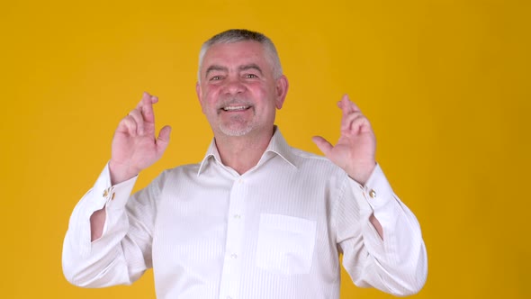 Elderly gray-haired man look at camera with crossed fingers wish isolated on yellow background