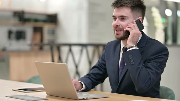 Young Businessman with Laptop Talking on Smartphone in Office  alt