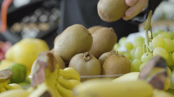 Close-up of Kiwi Fruits Lying on Shelf in Grocery As Unrecognizable Man Putting One Into Shopping alt