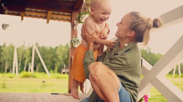 A Blond Baby Boy in Orange Shorts Laughing While His Mom Hugging and Kissing Him