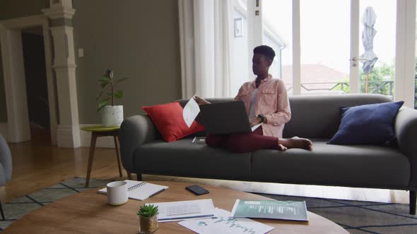 African american woman reading a document and using laptop while working from home alt