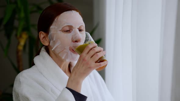 Mature Woman in Face Mask Drinking Vegetarian Smoothie Looking Out the Window Indoors alt