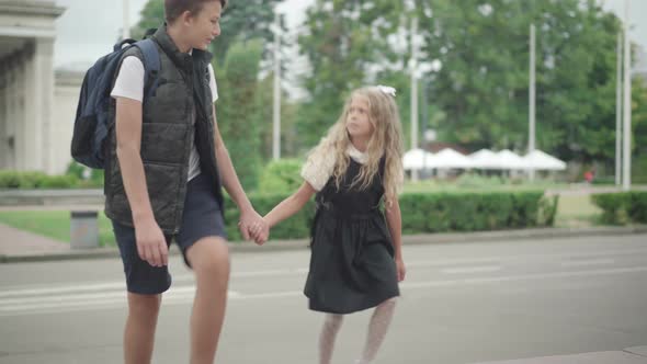 Pretty Girl Walking To School Holding Brother's Hand. Portrait of Little Cute Caucasian Schoolgirl alt