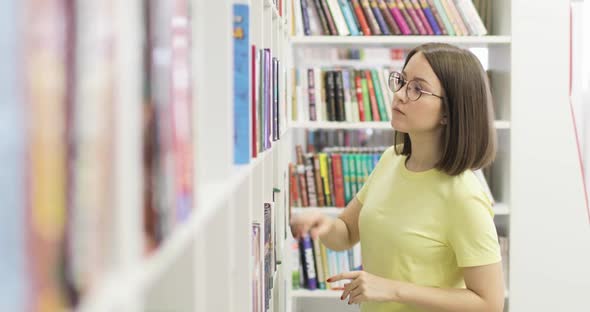 Young Beautiful European Female Student Wearing Glasses Chooses Book in College Library
