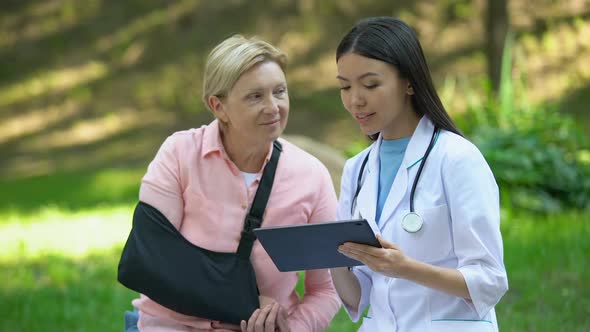 Asian Medical Worker With Tablet Smiling to Injured Senior Patient in Arm Sling alt