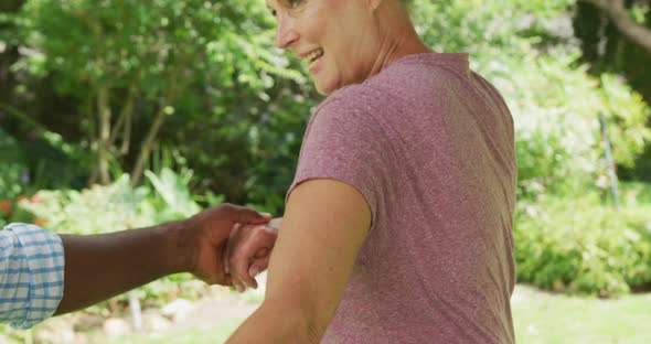 Happy senior diverse couple wearing shirts and dancing in garden alt