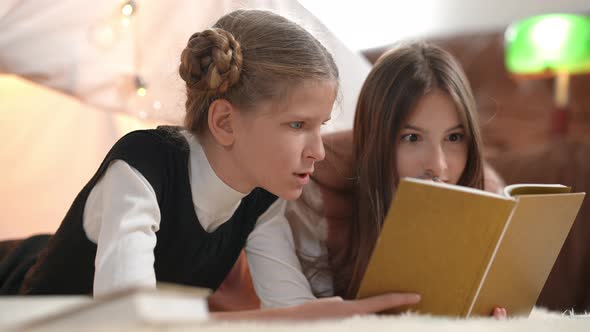Excited Absorbed Teenage Girls Reading Book Talking Lying Indoors at Tent in Living Room alt
