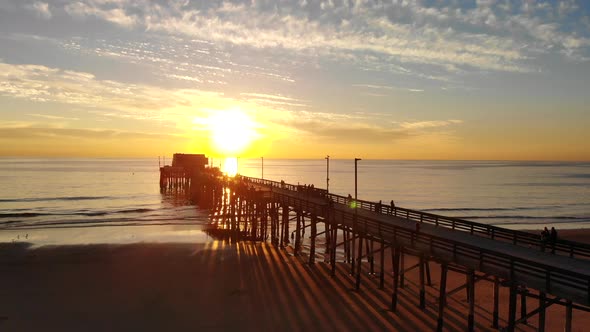 Drone rising over the Newport Beach pier at sunset over the pacific ocean as people walk along the C alt