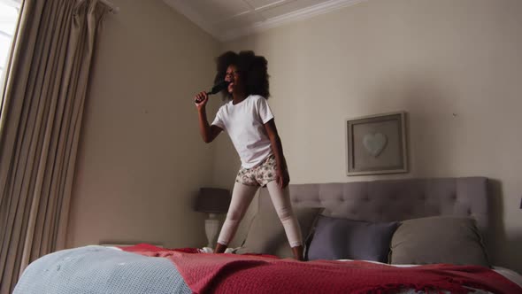 African american girl with hairbrush singing while standing on the bed at home alt
