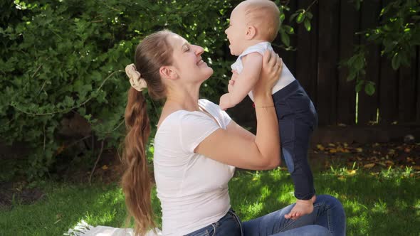Happy Cheerful Mother Holding and Lifting Up Her Little Baby Son in Backyard Garden alt