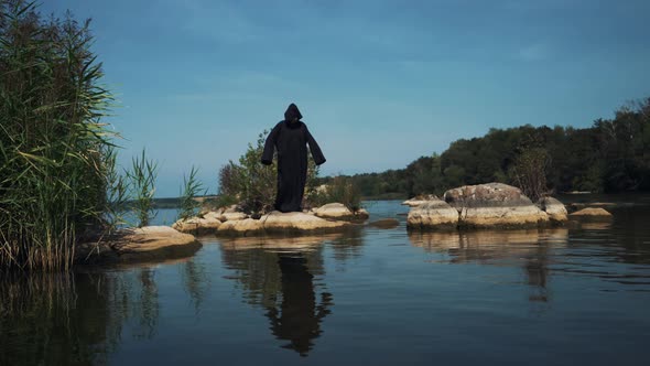 Evil Witch in Black Cloak with Hood on Stones Near the River alt